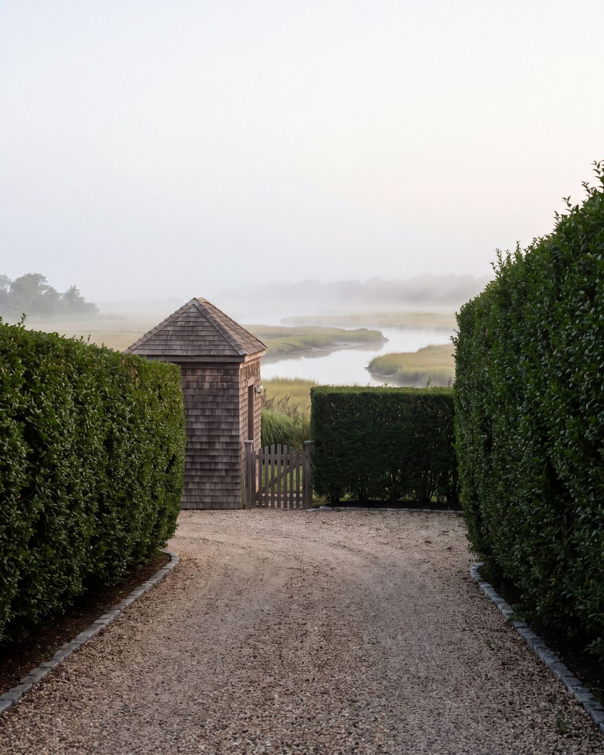 A pea-gravel lane between tall clipped privet hedges leading to a small shingled gatehouse, with marsh grasses and a tidal creek in the morning mist beyond